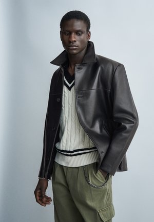 Νέος άνδρας που φοράει μαύρο δερμάτινο μπουφάν πάνω από λευκό πουλόβερ με πλεκτό σχέδιο και μαύρες ρίγες, χακί παντελόνι και ασημένιο αλυσιδάκι ως αξεσουάρ.