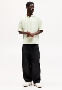 Lys grøn kortærmet polo med klassisk krave, parret med sorte vide jeans og hvide sneakers, stående på en neutral baggrund.