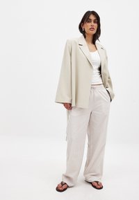 Svetlo bež oversize blazer, bela majica bez rukavov in široke hlače z kremastimi črtami. Obleko dopolnjujejo črne ravne sandale. Gladek material, sproščen kroj.
