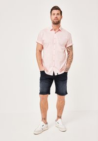 Kurzärmliges, rosa Hemd mit einer dezenten Musterung, kombiniert mit knielangen dunklen Jeansshorts und weißen Sneakern.