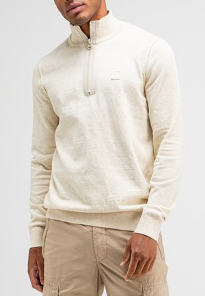 Beige ritsshirt met een hoge kraag, gemaakt van een gestructureerde gebreide stof. Beschikt over een klein logopatch op de borst en geribbelde manchetten.