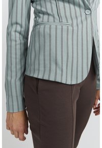 Világoskék csíkos zakó, slim fit szabású, egy gombos záródással és oldalsó zsebbel, barna szabott nadrággal párosítva.