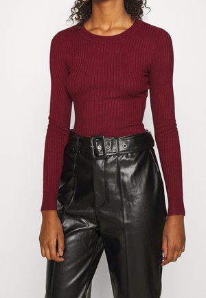 Femme portant un haut bordeaux ajusté à manches longues côtelé, rentré dans un pantalon en cuir noir taille haute avec une large ceinture et une grosse boucle.