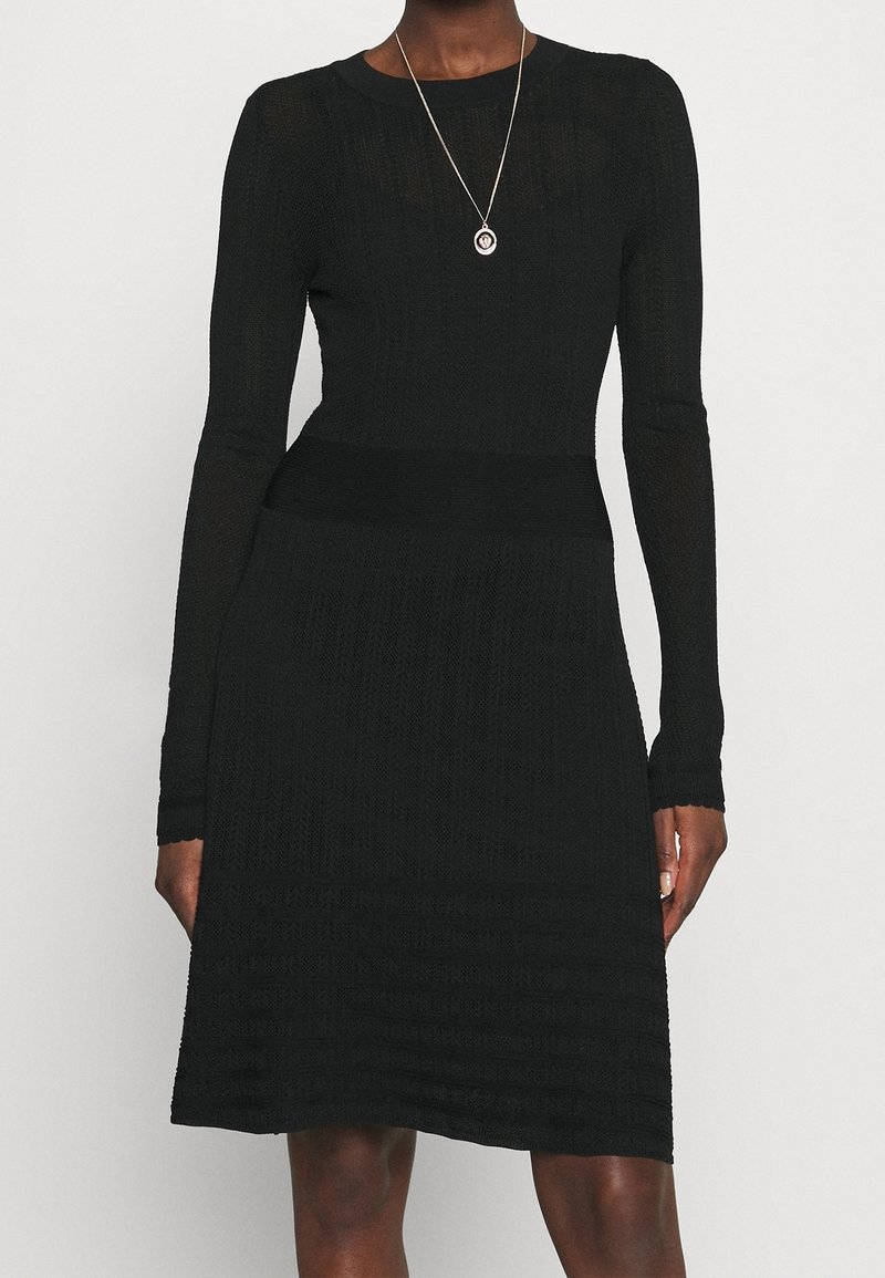 Robe en tricot noire à manches longues avec un haut ajusté, une jupe évasée et un motif en relief. Un collier avec un pendentif en argent est porté.