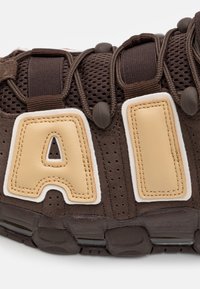 Brun atletiksko med en mesh-överkonstruktion, gummisulor och framtr ädande krämfärgade läderbokstäver "A" och "I" på sidan.