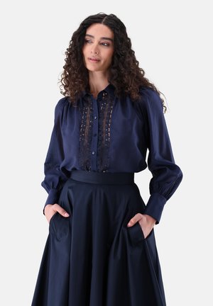 Donna con capelli ricci che indossa una camicetta blu navy con dettagli in pizzo e una gonna blu navy a vita alta, in piedi con le mani in tasca su sfondo bianco.