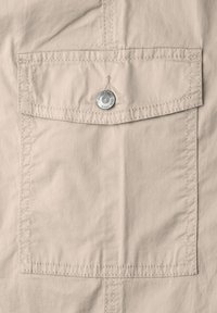 Poche en tissu beige avec rabat et un bouton rond en argent gravé de "S. Street One since 1989" sur un matériau cousu.