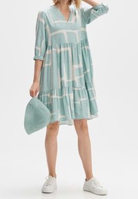 Vaaleansininen mekko valkoisilla abstrakteilla kuvioilla, V-pääntiellä ja kolme neljäsosaa hihoilla. Yhdistetty teksturoidun teal-värisen laukun ja valkoisten tennareiden kanssa.