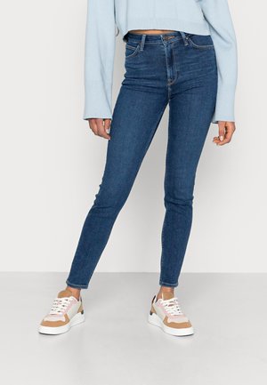 Personne portant un jean skinny bleu foncé, un haut à manches longues bleu clair, et des baskets blanches, beiges et roses, debout sur un sol clair.