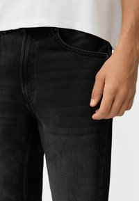 Vaqueros de mezclilla negros con corte slim, que presentan una textura sutil, bolsillos laterales y cierre con botón, combinados con una camiseta blanca.