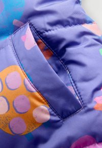 Lila mintás anyag, amely narancssárga és rózsaszín kör alakú formákat ábrázol, egy kis zseb van varrva az anyagba. Simább tapintású.