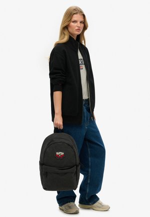 Chaqueta negra con cierre sobre una camiseta gris, jeans de pierna ancha azules, zapatillas grises y una mochila negra con un parche de logo rojo y blanco.