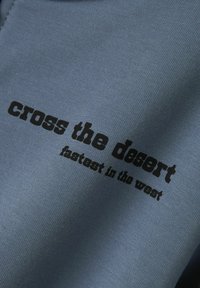 Zils kokvilnas audums ar gludu tekstūru, kurā ir melns uzraksts: "šķērso tuksnesi" un "ātrākais Rietumos" treknā fontā.