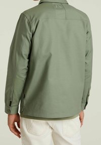 Olivengroene jas van een zachte, gladde stof met een kraaglijn, knoopmanchetten en een zijnaadlabel; gedragen over een licht shirt.