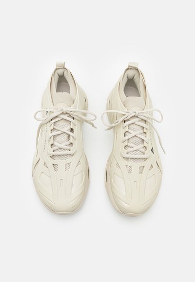 Beige athletic shoes with a mesh and synthetic upper, round laces, and reinforced sides. Features minimalistic design with ventilation details.