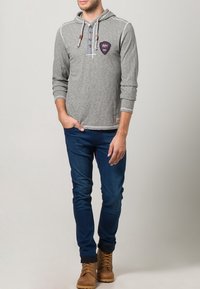 Grauer Hoodie mit strukturierter Optik, ausgestattet mit einer Fronttasche, Knopfverschluss und einem Logopatch. Kombiniert mit dunkelblauen Jeans und braunen Stiefeln.