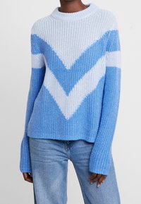 Pull tricoté en bleu clair et blanc avec un motif chevron. Coupe ample avec un col montant et des poignets côtelés. Texture douce.