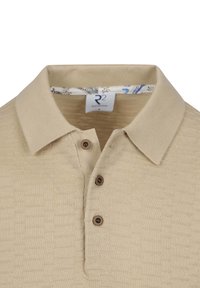 Beige poloshirt met een getextureerd patroon, drie bruine knopen en een kraag. Bevat een merklabel aan de binnenkant van de kraag.