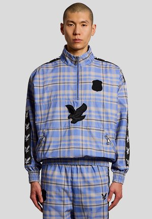 Man draagt een blauw-beige geruit trainingspak met zwarte arendpatches en een halve rits trui, staand tegen een effen achtergrond.