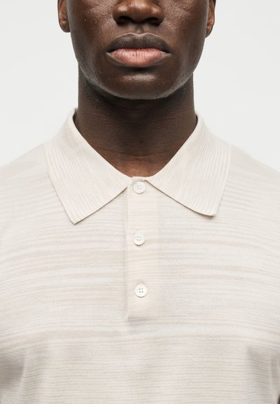 Κοντινό πλάνο ενός ατόμου που φορεί ένα ανοιχτό μπεζ πόλο πουκάμισο με μυτερό γιακά και τρία λευκά κουμπιά κλεισμένα.