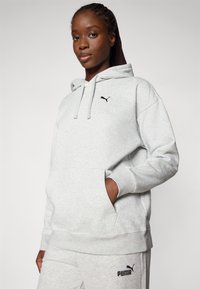 Svijetlosiva hoodie s kenguruh džepom, podesivim trakama i crnim Puma logotipom na prsima. Mekana, udobna tkanina.