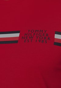 Červené bavlněné tričko s centrálním grafickým motivem v černých, bílých a námořnicky modrých pruzích, s textem "TOMMY HILFIGER NEW YORK EST 1985" v černé barvě.