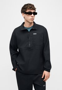 Νεαρός άνδρας που φοράει μαύρο μπουφάν Columbia με ημιφερμουάρ, ταιριαστά παντελόνια, μπλε πουκάμισο από κάτω και μαύρα ορθογώνια γυαλιά ηλίου, όρθιος με τα χέρια στις τσέπες.