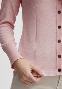 Svetlo roza pletena jakna z dolgimi rokavi, z gumbnim zapiranjem spredaj in rebrastimi manšetami. Gladka tekstura z diskretnimi vzorčnimi podrobnostmi.