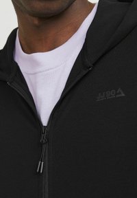 Schwarze Zip-Hoodie mit glatter Textur, mit einem Reißverschluss und einem kleinen Logo. Sie ist über einem helllila Shirt getragen.