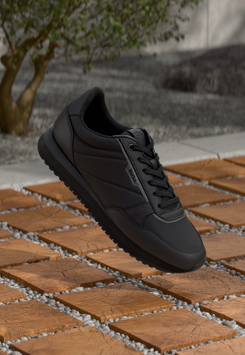 Baskets noires à lacets avec semelle texturée, flottant au-dessus d'un carreau en bois carré et de petits cailloux en extérieur.