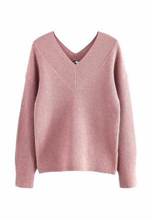Femme portant un pull côtelé rose, un jean bleu clair avec de petits motifs floraux et des baskets argentées métalliques, debout devant un fond vert.