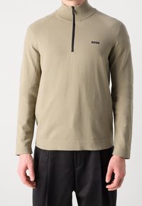 Beige långärmad tröja med svart fyrdelsdragkedja och "BOSS"-logotyp på bröstet, bärs tillsammans med svarta byxor, manlig modell beskuren vid nacken.