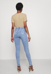 Woman with short curly hair wearing a striped t-shirt, light blue ripped skinny jeans, and olive green high-heeled sandals, facing away.