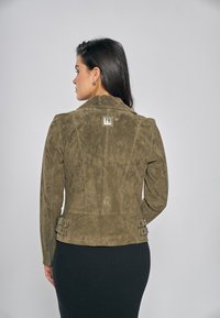 Olivgrön mockajacka med figurnära silhuett, bältesdetaljer i midjan och en synlig logotyp på övre delen av ryggen.