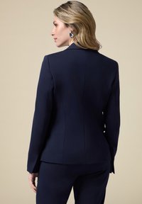 Blazer azul marino con un corte estructurado, textura suave, solapas de muesca y sutiles detalles de costura, acompañado de pantalones a juego.