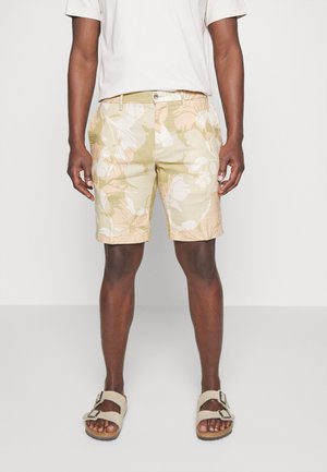Mand iført beige shorts med blomsterprint, hvid t-shirt og beige sandaler med spænde, stående foran en ensfarvet baggrund.
