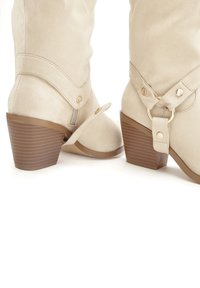 Botas de tobillo de ante beige con tacón apilado, que cuentan con una correa con un anillo dorado y detalles de botón. Textura suave y diseño de punta redonda.