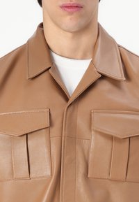 Kohvane nahkshirt jakk, millel on kaks esiflakki taskut, lai krae ja sile tekstuur. Voltitud kraed ja korrektsed õmbluse detailid.
