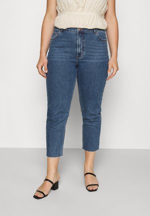 Nainen, jolla on päällään siniset korkeavyötäröiset lyhytlahkeiset farkut raakana helmalla, beigen värinen hihaton paita ja mustat avoimet korkokengät, seisomassa harmaalla lattialla.