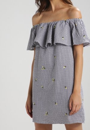 Kvinde iført off-shoulder sort-hvid ternet kjole med små gule blomsterbroderier, stående mod en ensfarvet lys baggrund.