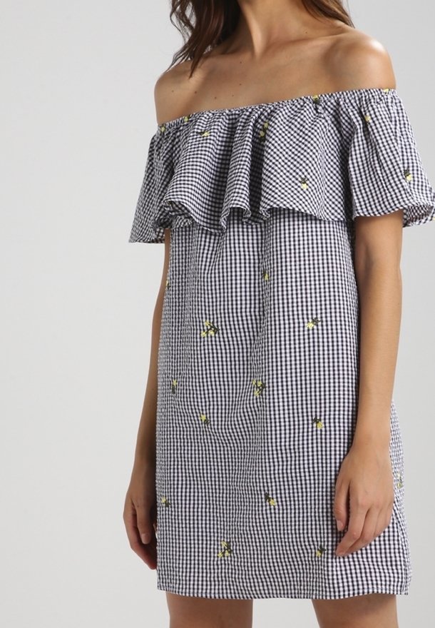 Femme portant une robe à carreaux vichy noir et blanc à épaules dénudées avec une petite broderie florale jaune, se tenant devant un fond clair uni.