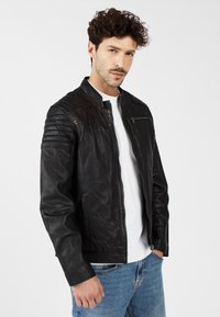 Zwarte leren jas met gevoerde schouderdetails, aansluitend ontwerp, vooraan ritsluiting en zijzakken. Draag bij een wit shirt en blauwe jeans.