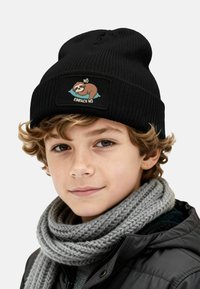 Black knitted beanie with a sloth graphic patch featuring the text "NÖ EINFRACH NÖ." Worn with a gray knitted scarf and dark jacket.