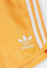 Pantaloni sportivi gialli realizzati in tessuto liscio. Presentano tre strisce bianche lungo il lato e un logo Adidas bianco.