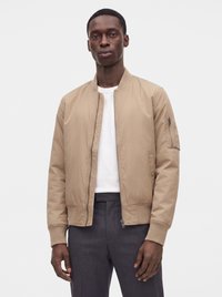 Homme portant une veste bomber beige par-dessus une chemise blanche et un pantalon foncé, debout devant un fond uni gris clair.