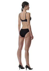 Vrouw staande in een zwarte beha, zwarte onderbroek en zwarte hakken, gezien van achteren tegen een witte achtergrond.