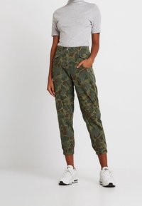 Camo cargobroeken in gedempte groene tinten met elastische cuffs, zijzakken en een relaxed fit, gecombineerd met een lichtgrijze T-shirt met korte mouwen.