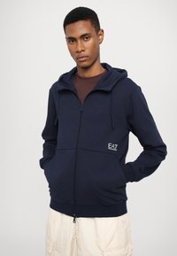Tamsiai mėlynas zip-up džemperis su reguliuojama kapuce, priekinėmis kišenėmis ir EA7 logotipu. Pagamintas iš minkštos medžiagos su glotnia tekstūra.