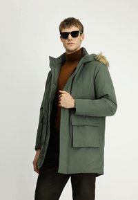 Mężczyzna w czarnych okularach przeciwsłonecznych, zielonej kurtce z kapturem wykończonym futrem, brązowym swetrze z golfem i ciemnych spodniach, stojący na jednolitym tle.