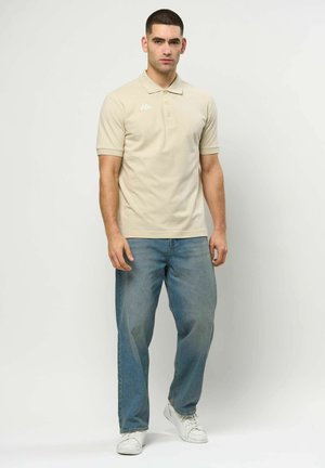 Moški v bež polo majici s kratkimi rokavi, svetlo modrih jeansa in belih supergah stoji ob enobarvnem svetlem ozadju.
